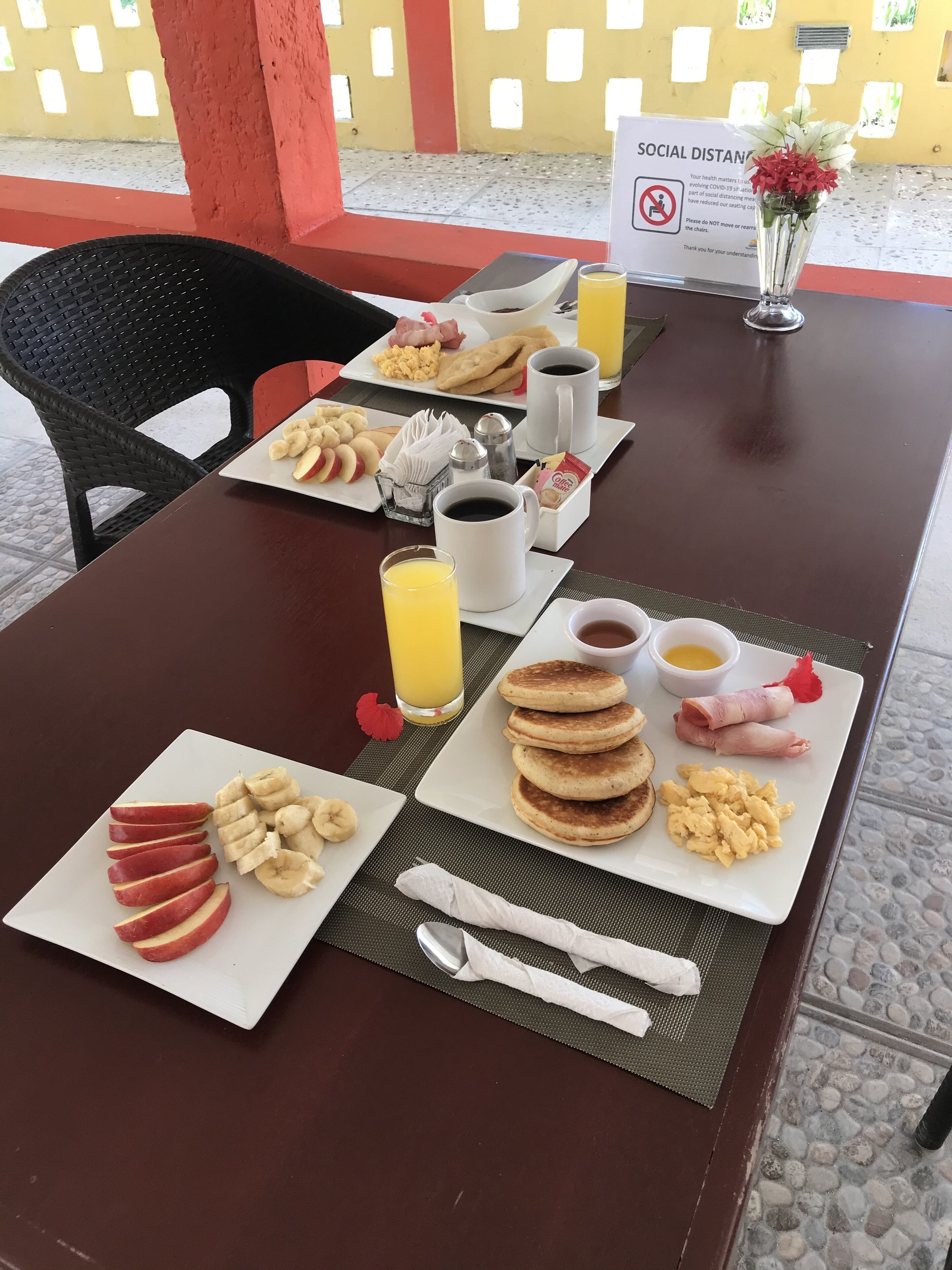 Mesa de café da manhã com pratos de comida, incluindo panquecas, ovos e frutas, acompanhados de xícaras de café e suco.