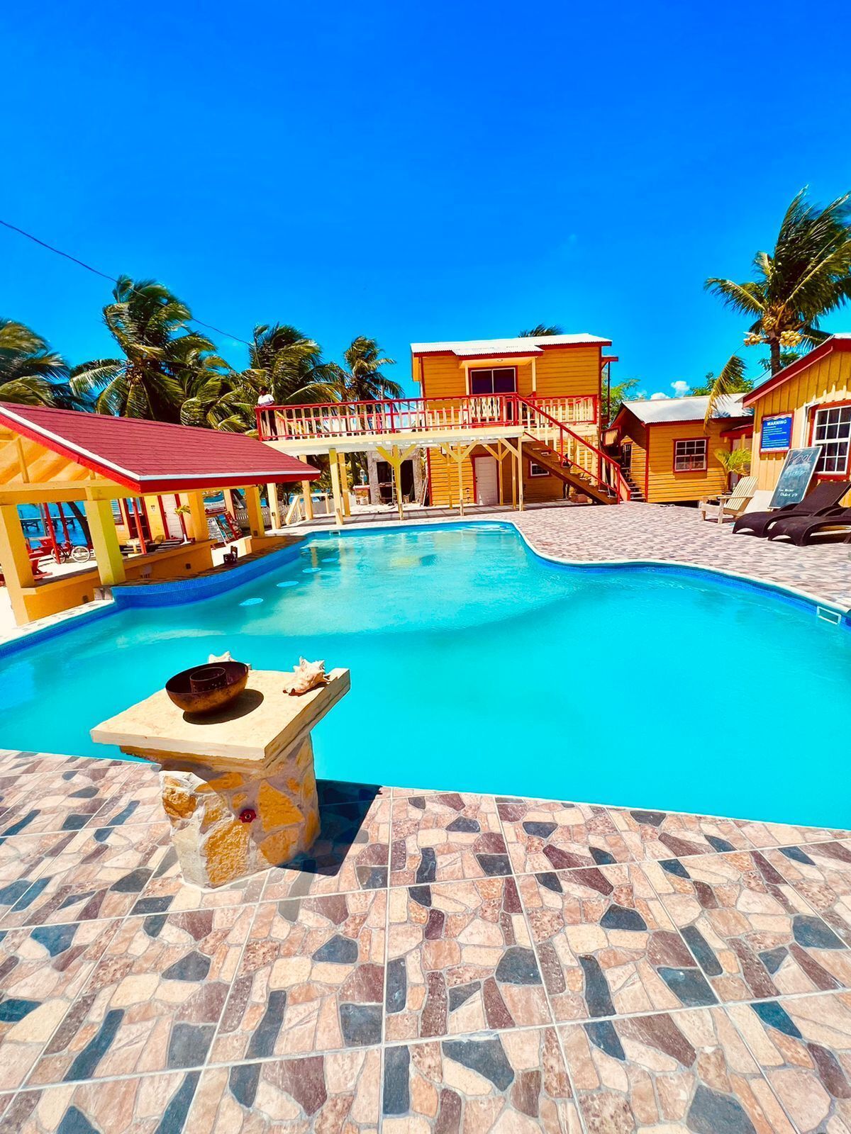 A swimming pool with a tiled deck, a covered seating area, and a building with a red roof.