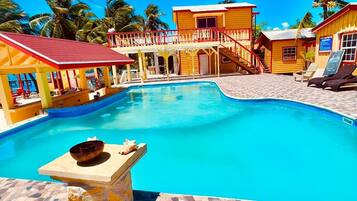 A swimming pool with a tiled deck, a covered seating area, and a building with a red roof.