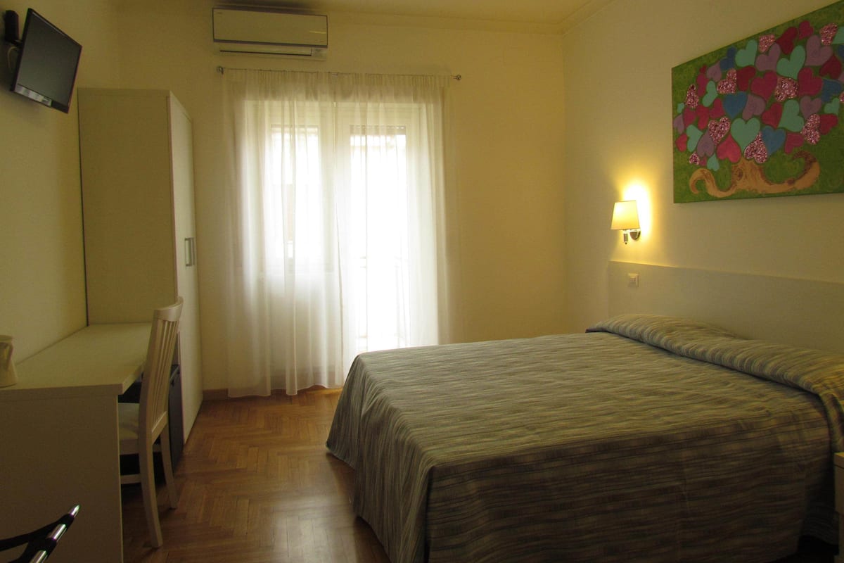 A hotel room with a bed, a desk, a chair, a television, and a painting on the wall.