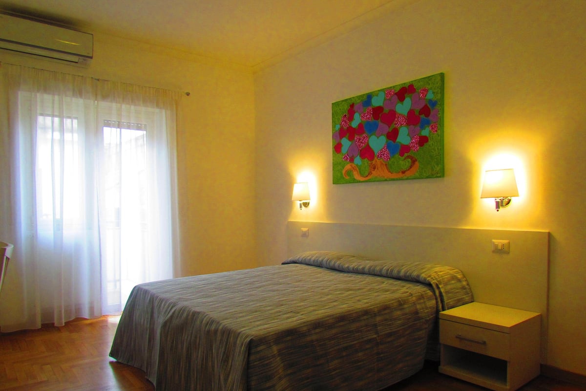 A hotel room with a bed, bedside tables, a wall-mounted lamp, and a colorful artwork.