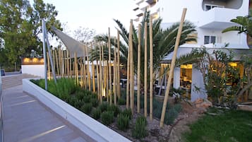 Un edificio moderno con fachada blanca, rodeado de un jardín ajardinado con bambú y palmeras.