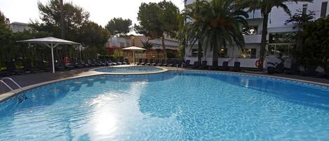 A swimming pool with lounge chairs and umbrellas, surrounded by palm trees and a multi-story building.