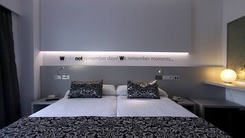 Una habitación de hotel moderna con una cama grande, dos almohadones decorativos, una mesita de noche con lámpara y una lámpara de pared.