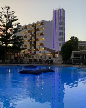 Een hotel met een zwembad, ligbedden en een hoog gebouw met verlichte strepen.