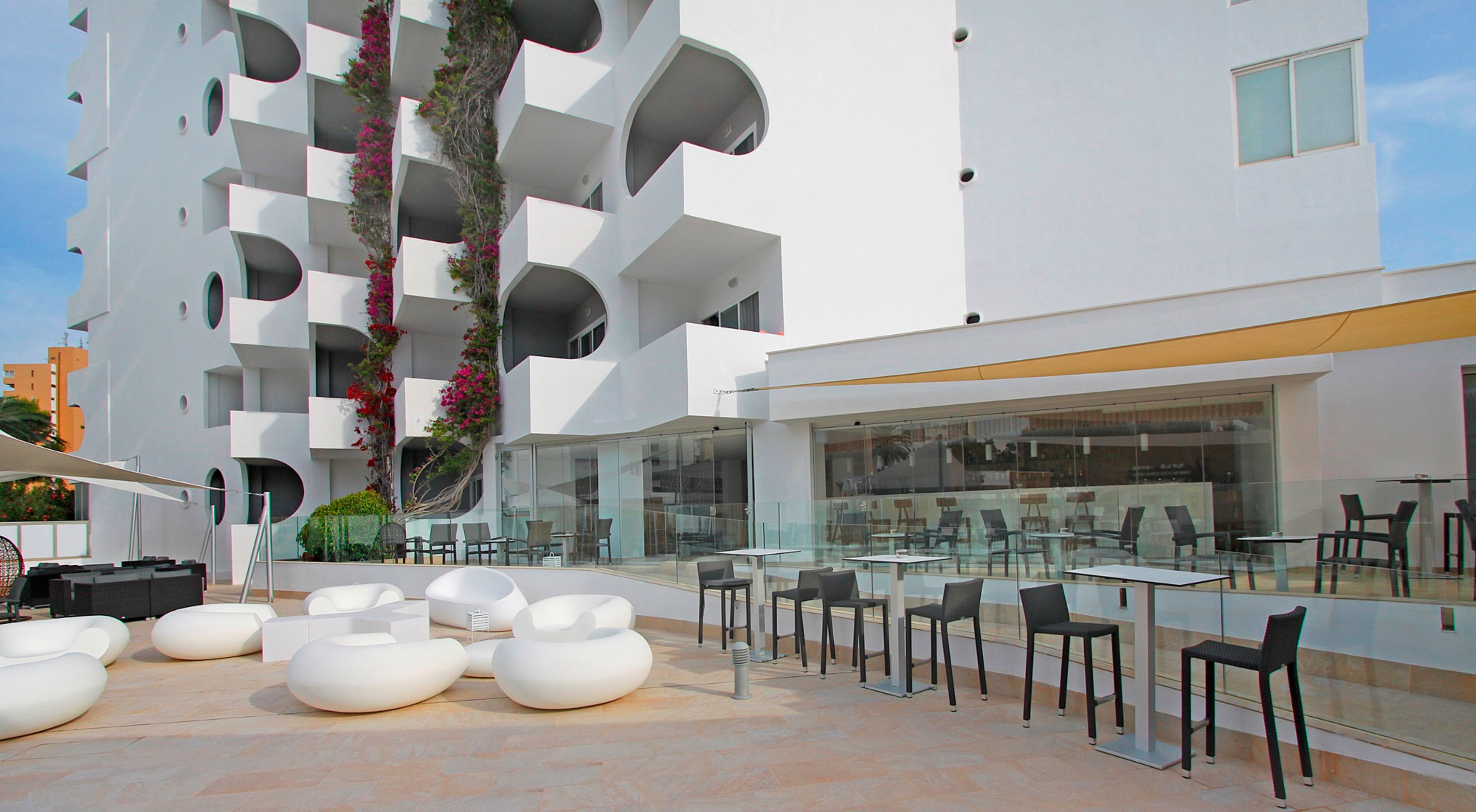 A modern outdoor seating area with white planters and tables, surrounded by a white building with balconies and a glass facade.