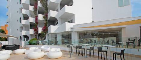 A modern outdoor seating area with white planters and tables, surrounded by a white building with balconies and a glass facade.