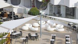Un área de estar al aire libre con sillas y mesas modernas, rodeada de sombrillas blancas y una barandilla de cristal.