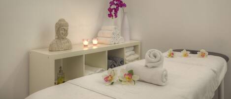 A neatly arranged spa room with a bed, a shelf with towels, a bust sculpture, candles, and flowers.