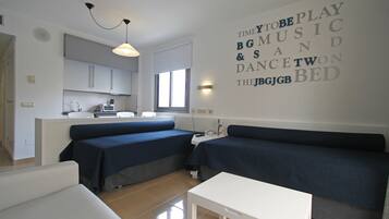 Una sala de estar moderna con un sofá, una mesita blanca y una zona de cocina con armarios y una ventana.