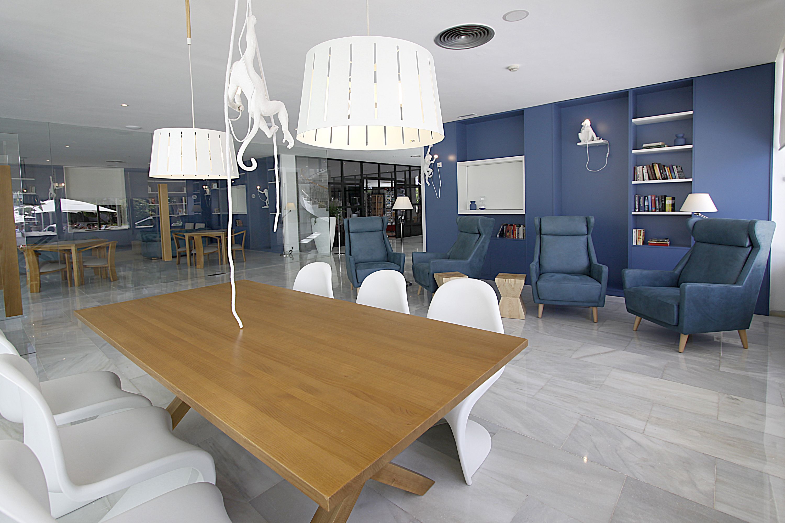 Ein modernes Wohnzimmer mit einem Holztisch, weißen Stühlen, blauen Sesseln und einem Bücherregal.