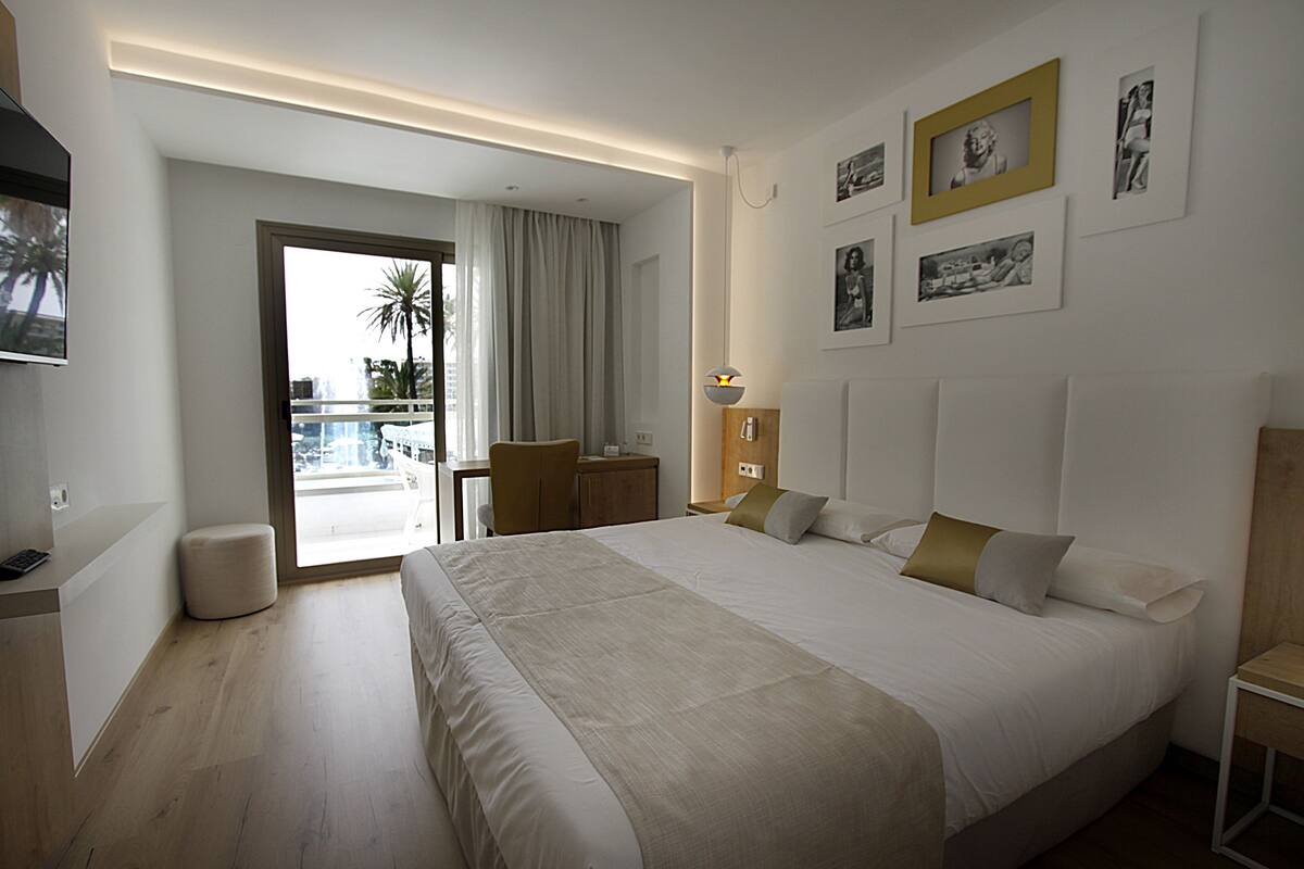 A hotel room with a large bed, a desk, a chair, a small stool, a TV, and a balcony with a view of palm trees and buildings.