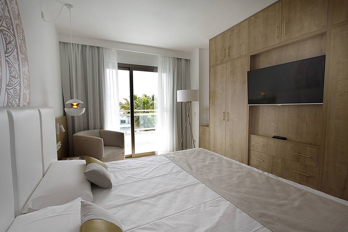 A modern hotel room with a large bed, a TV mounted on the wall, a desk with a lamp, and a view of the outdoors through the window.
