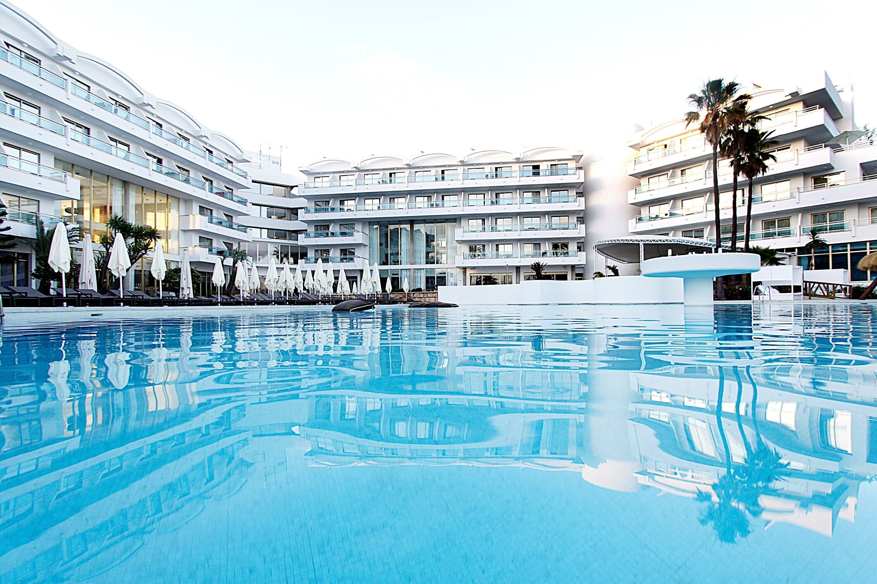 A large indoor swimming pool with multiple levels of balconies, white umbrellas, and palm trees.