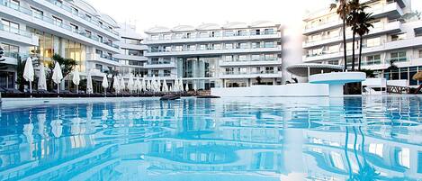 A large indoor swimming pool with multiple levels of balconies, white umbrellas, and palm trees.