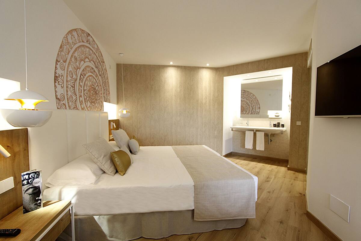 A modern hotel room with a large bed, bedside lamps, a wooden floor, a bathroom with a mirror and sink, and a TV mounted on the wall.