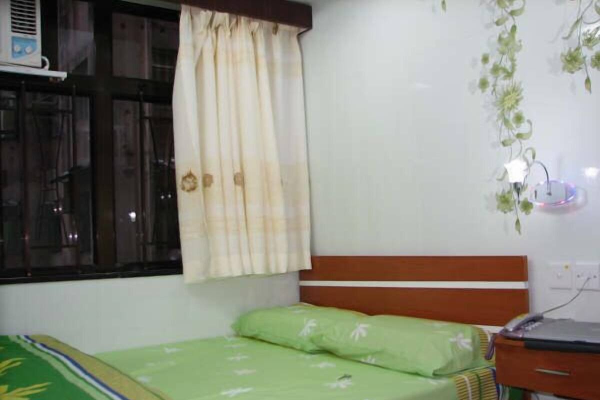 A small, single-bed room with a wooden headboard, a green bedspread with floral patterns, a beige curtain, and a wall-mounted air conditioner.