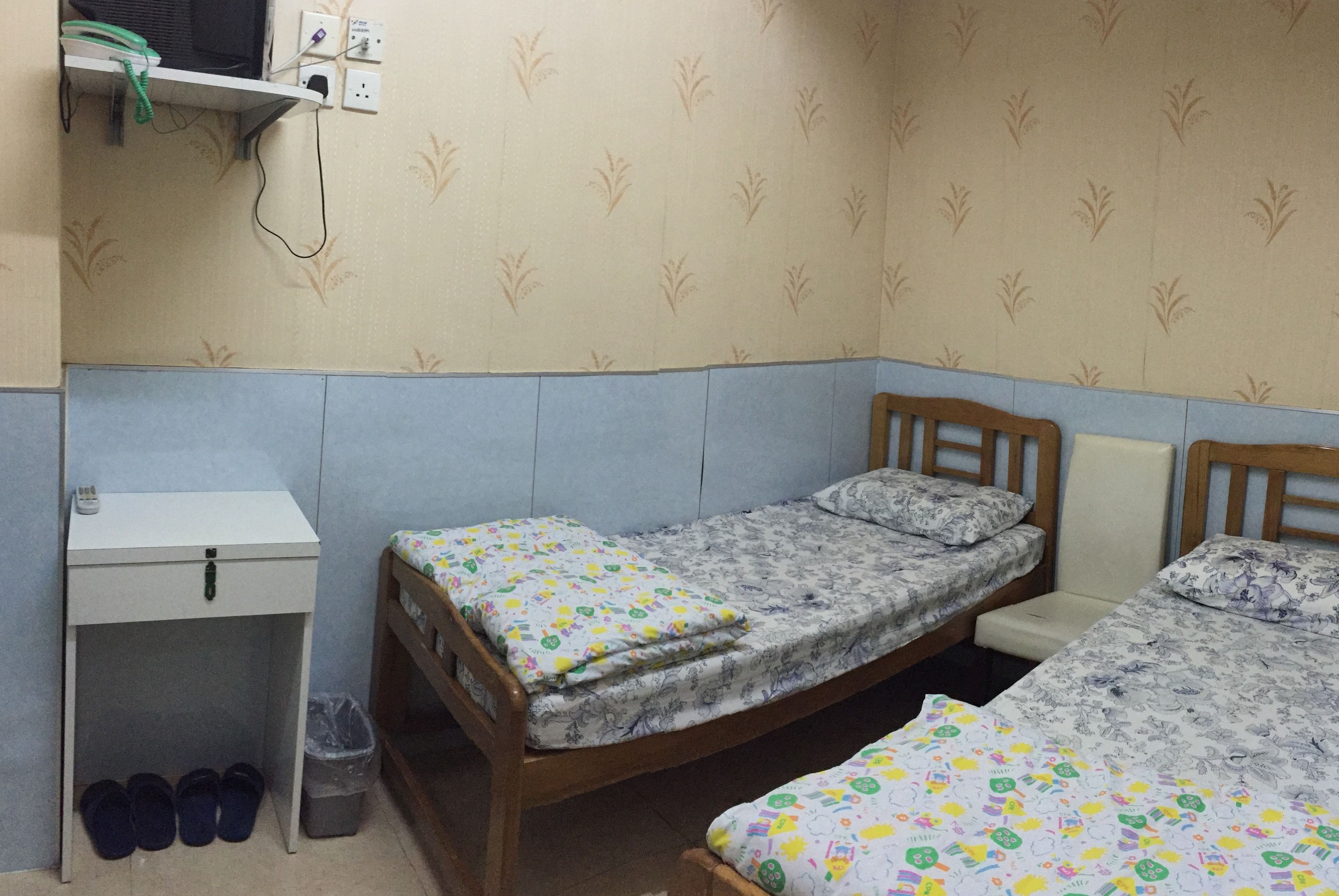 A small, single-room accommodation with two beds, a bedside table, a small desk, and a shelf with a power outlet.