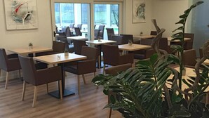 Un espace repas moderne avec des tables et des chaises en bois, de grandes fenêtres et des plantes d’intérieur.