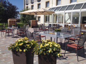 Une terrasse extérieure avec des tables et des chaises, un grand parasol et des plantes en pot.