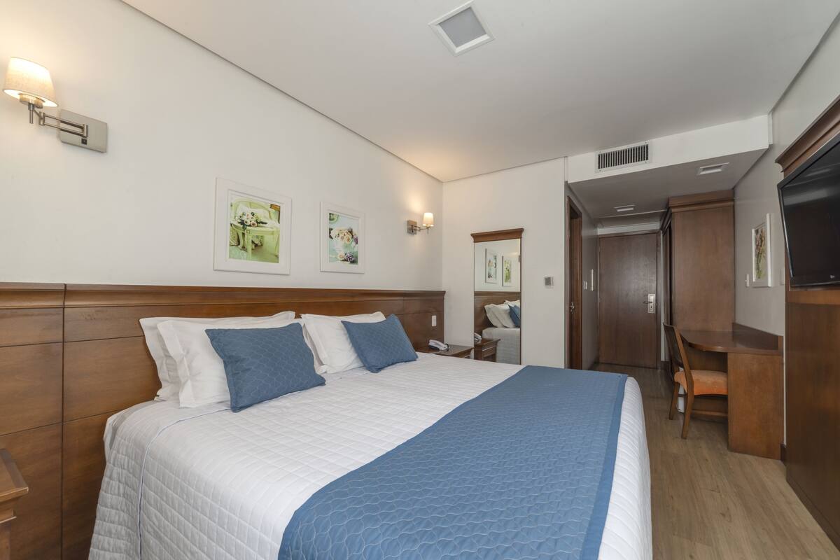 Una habitación de hotel con una cama grande, cabecero de madera, lámparas fijadas a la pared y un escritorio con una silla.