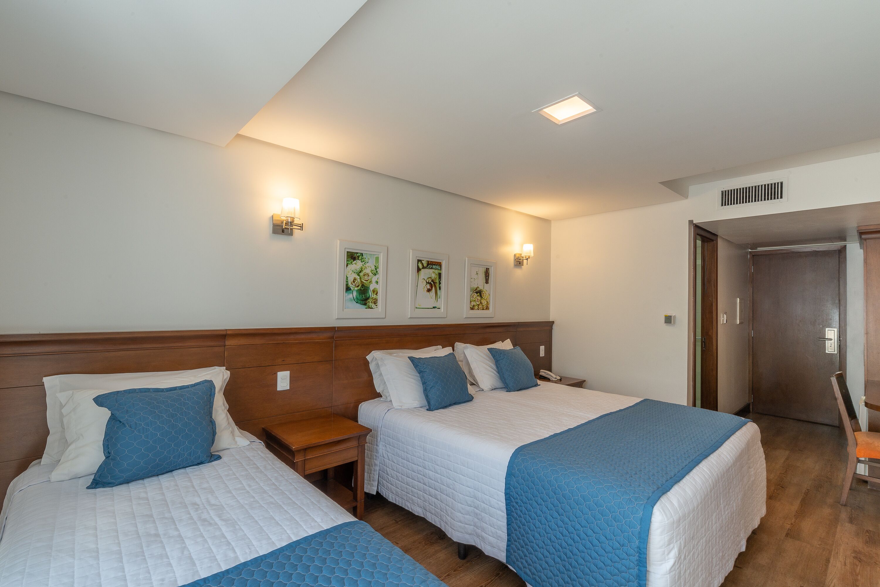 Une chambre d’hôtel avec deux lits, une tête de lit en bois, des tables de chevet, des lampes fixées au mur et des œuvres d’art encadrées accrochées au mur.