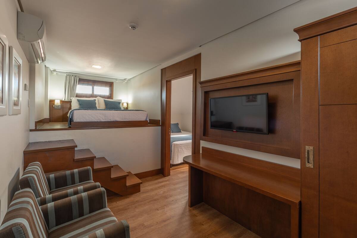 Una habitación compacta con una cama, un televisor fijado a un mueble de madera, una escalera que conduce a un nivel superior y una zona de estar con sofás a rayas.