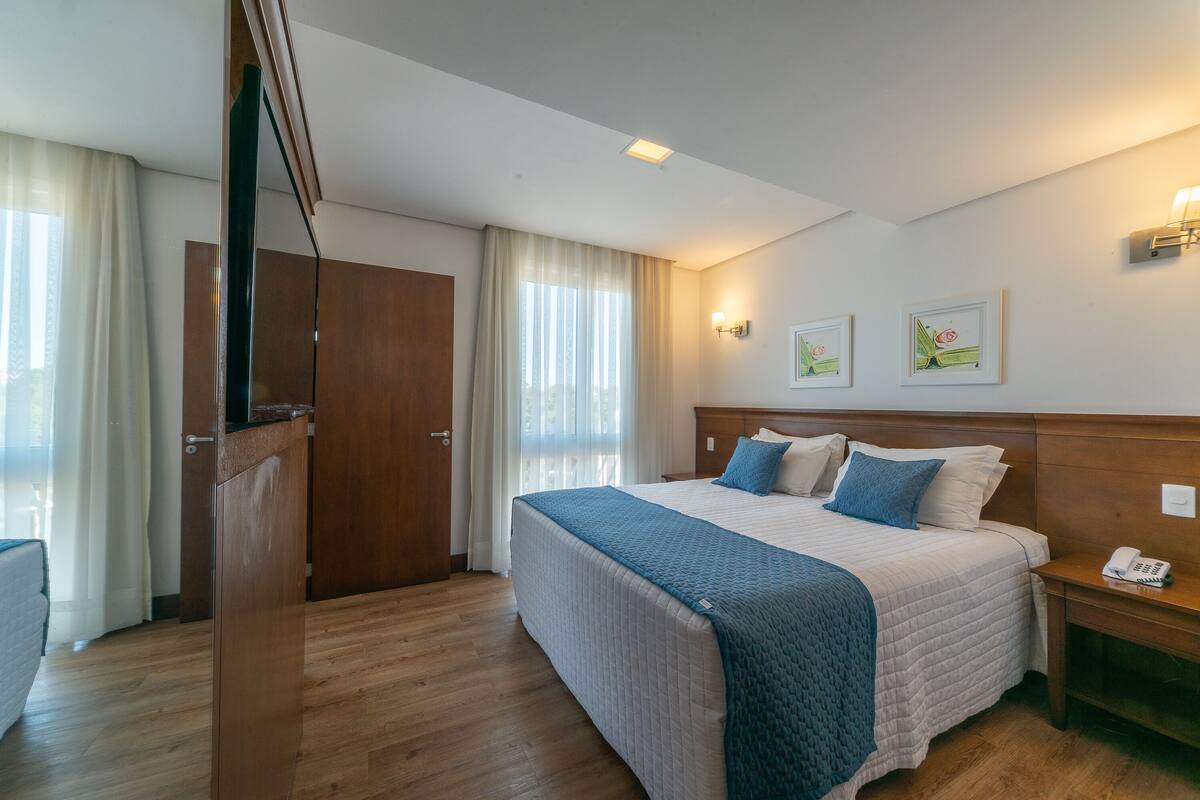 Una habitación de hotel con una cama grande, un cabecero de madera, una mesita de noche con un teléfono y una ventana con cortinas.