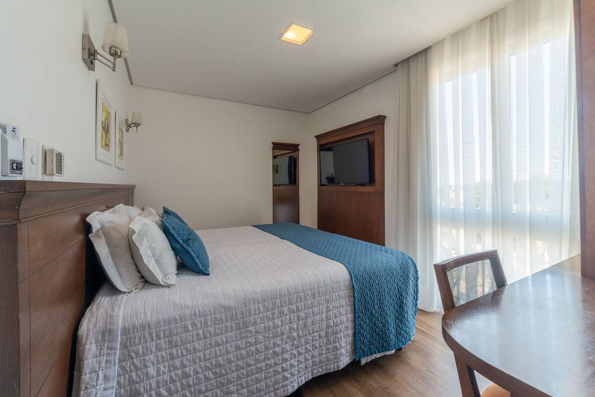 Un dormitorio con cama, cabecera de madera, mesita de noche, silla y ventana con cortinas.