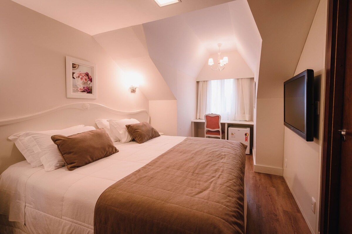 A bedroom with a bed, a desk, a chair, a television, and a window with curtains.