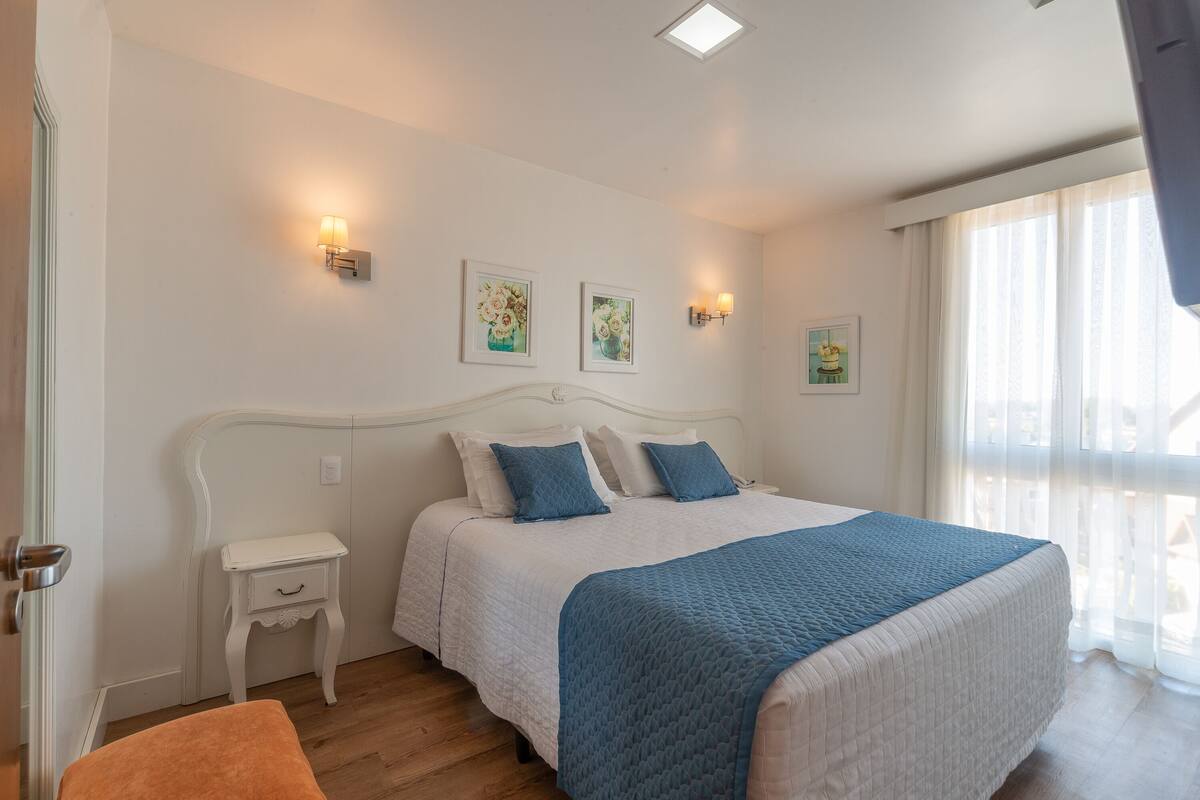 Un dormitorio con una cama blanca, almohadas azules y una colcha acolchada azul. Hay dos lámparas de pared y tres cuadros enmarcados.
