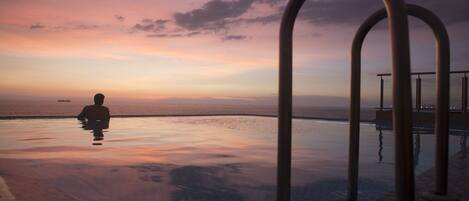 Una piscina infinita tranquila con una persona en el agua, con vista a la puesta de sol sobre el océano.