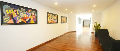 A hallway with wooden flooring, three framed artworks on the wall, and a black chair near a potted plant.
