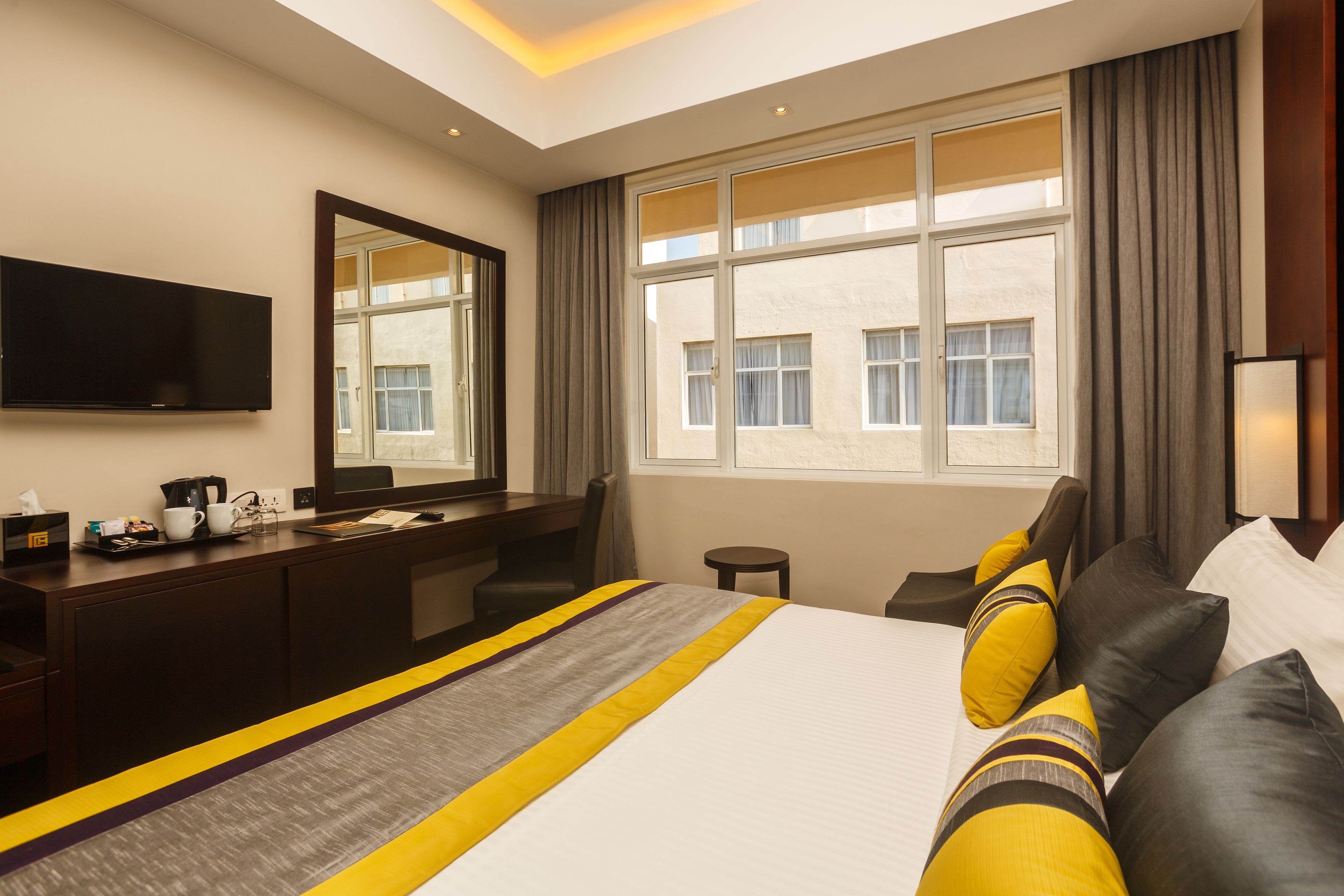 A hotel room with a large bed, a desk with a flat-screen TV, a mirror, a window with curtains, and a view of buildings outside.