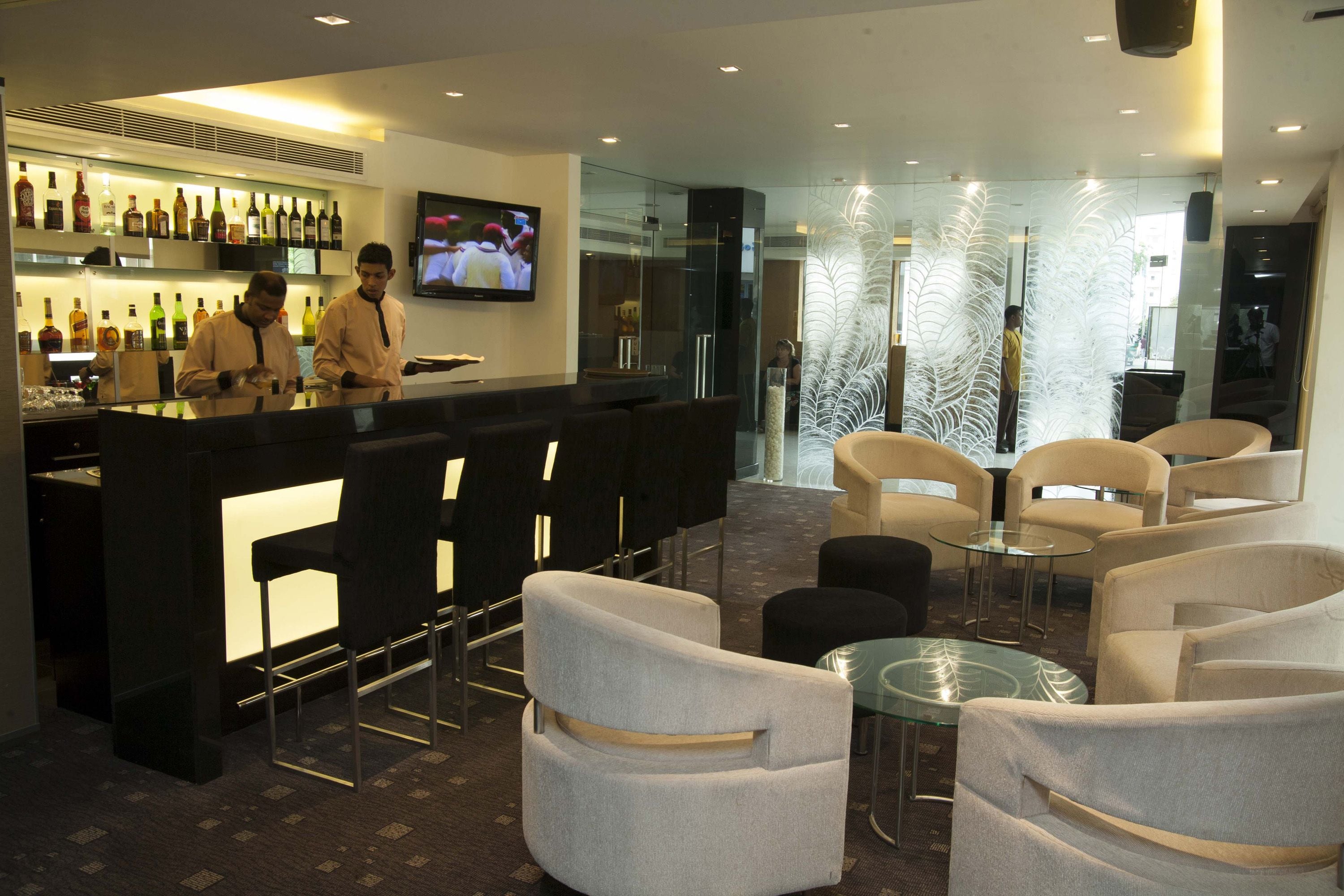 Área de bar moderna com balcão, banquetas e uma tela de televisão. Há divisórias de vidro e um elemento decorativo na parede.