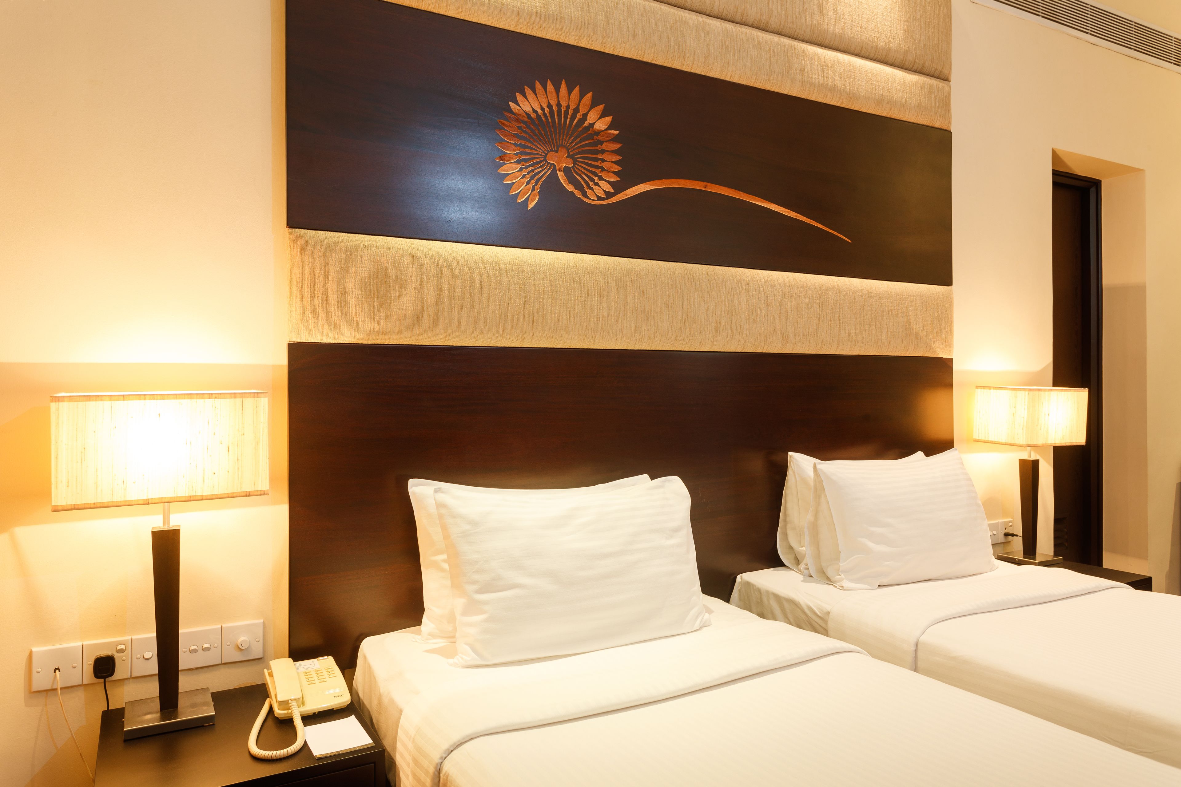 A hotel room with two beds, a wooden headboard with a floral design, bedside lamps, a telephone, and a power outlet.
