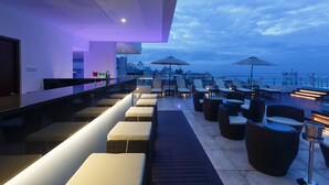 Um bar no terraço com assentos e guarda-sóis, com vista para uma paisagem urbana ao anoitecer.