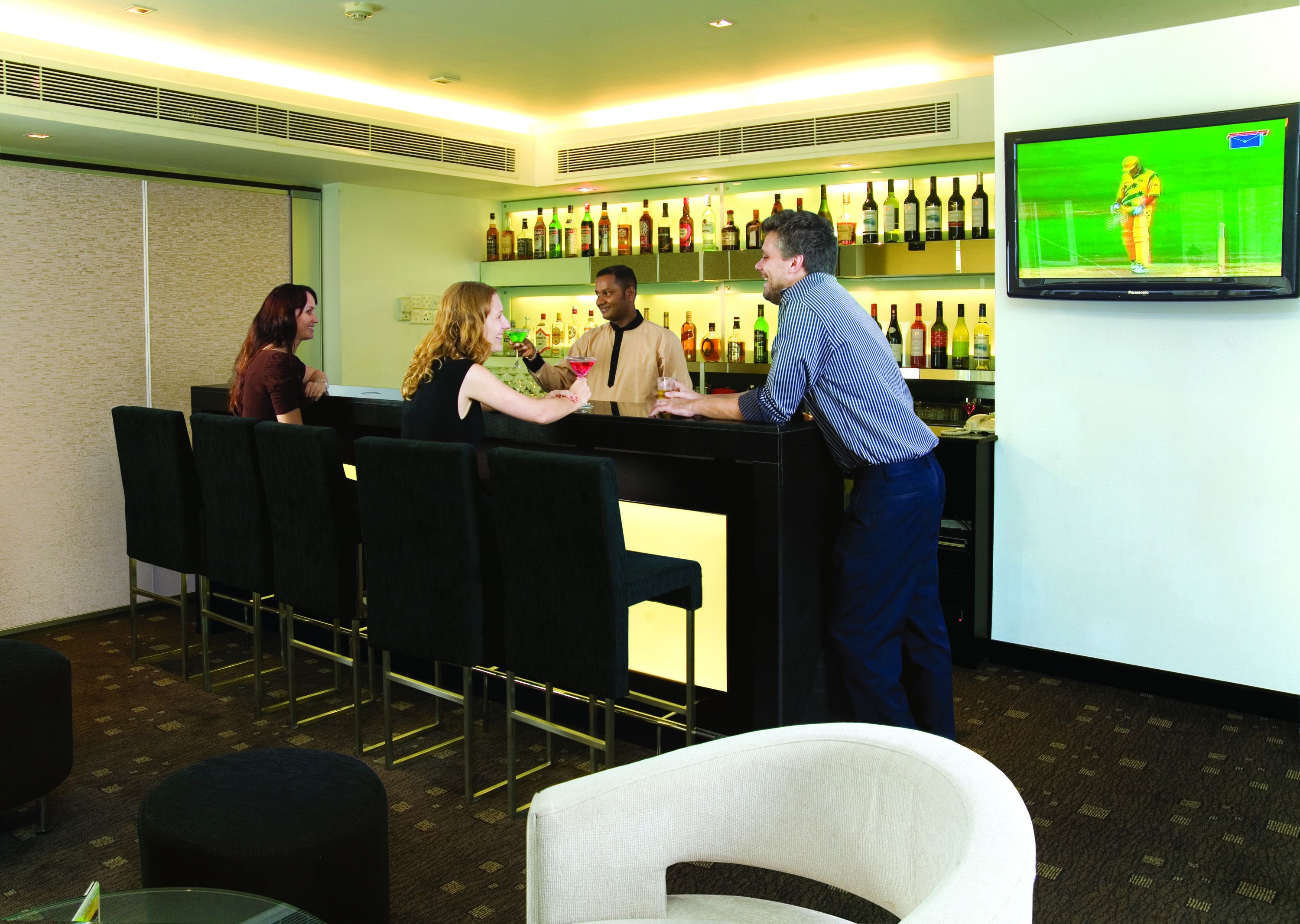 A bar area with a television screen displaying a cricket match, a bar counter, and several chairs.