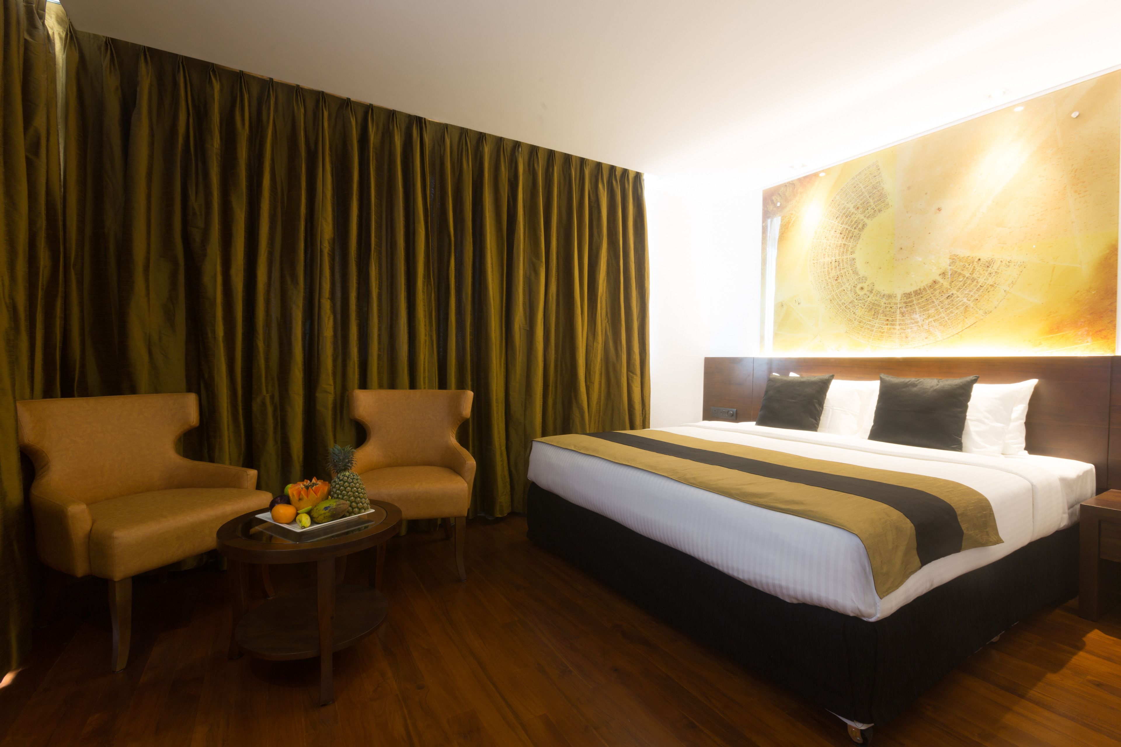 Ein Hotelzimmer mit einem Bett, zwei Sesseln, einem kleinen Tisch mit Früchten und einem großen abstrakten Gemälde an der Wand.