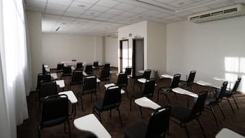 Un salón de conferencias con filas de sillas negras y mesas blancas, una pizarra y un equipo de aire acondicionado en el techo.