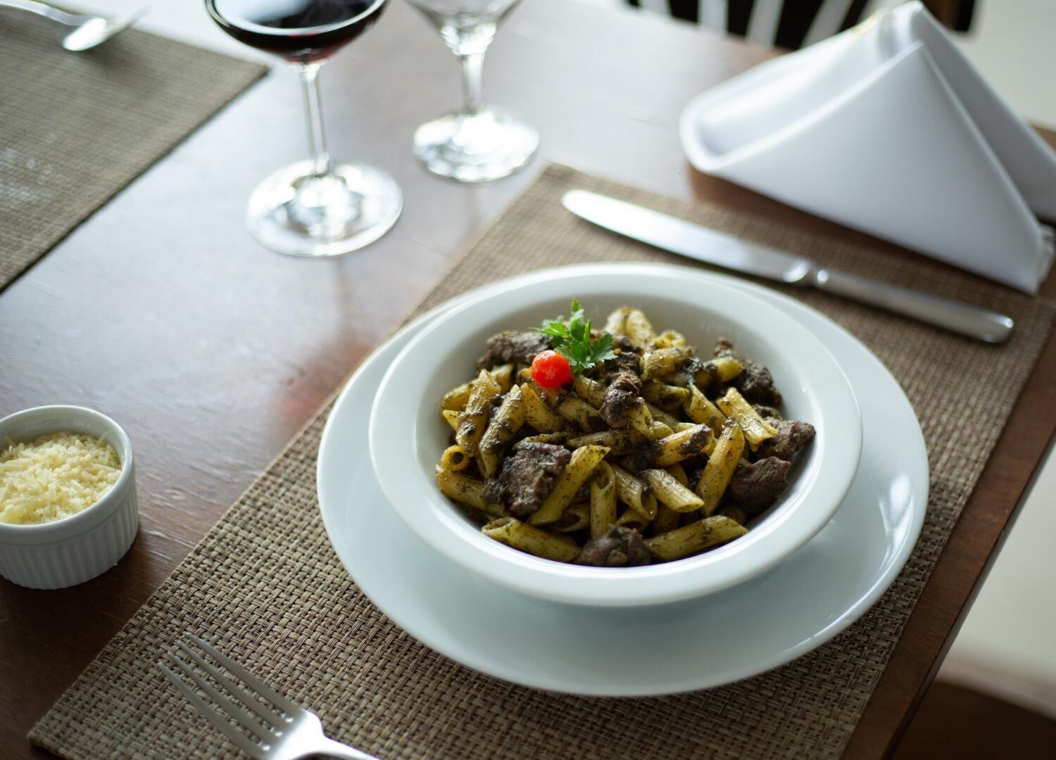 Un plato de pasta con carne y una decoración de tomate cherry, acompañado de un tenedor y un cuenco pequeño de queso rallado.