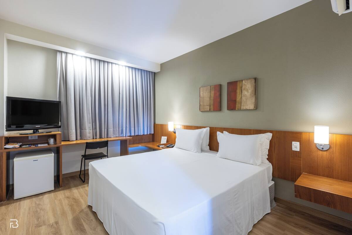 Una habitación de hotel con una cama grande, televisión, escritorio y un refrigerador pequeño.