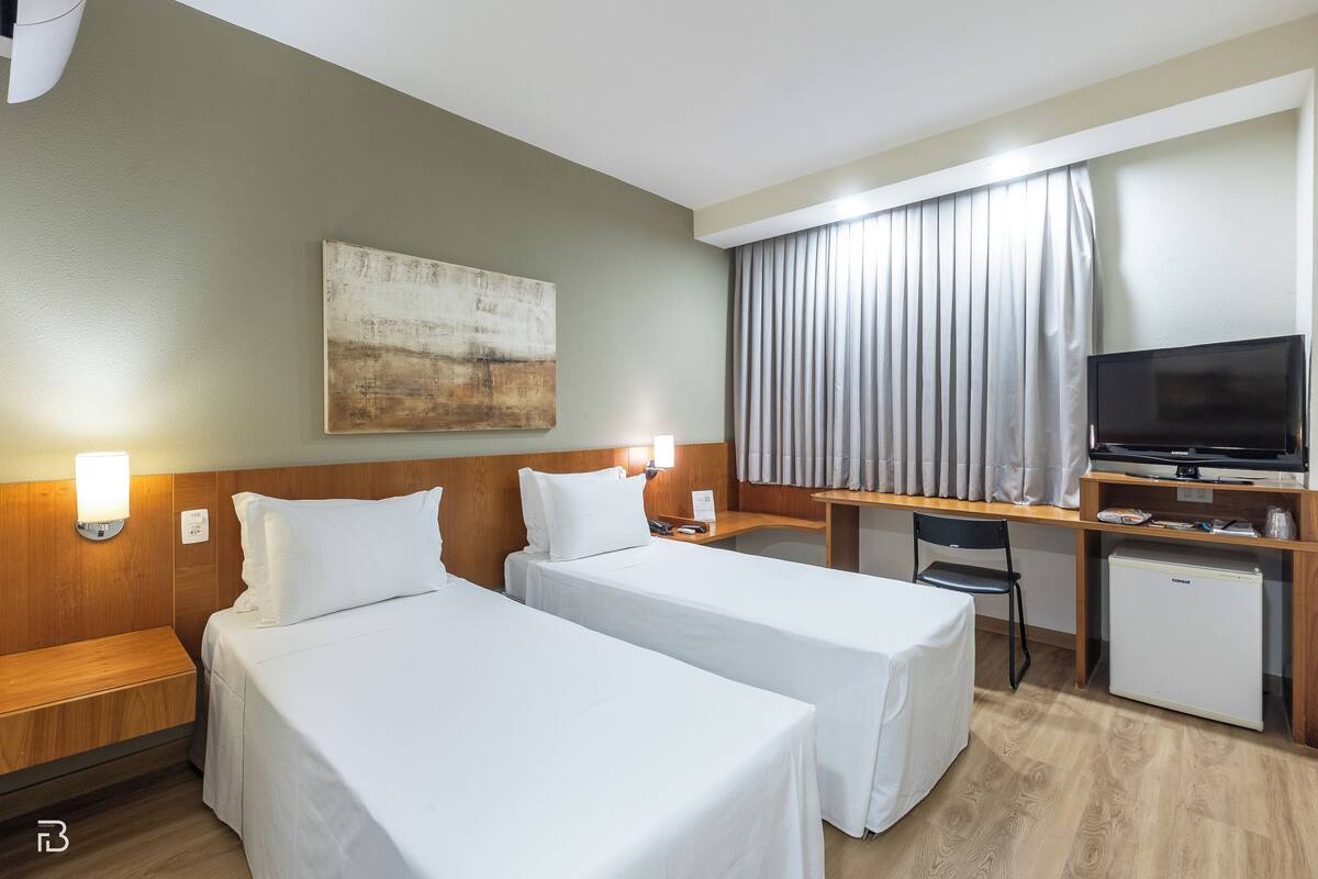 Una habitación de hotel con dos camas individuales, un escritorio con televisor, un refrigerador pequeño y un ventanal con cortinas.
