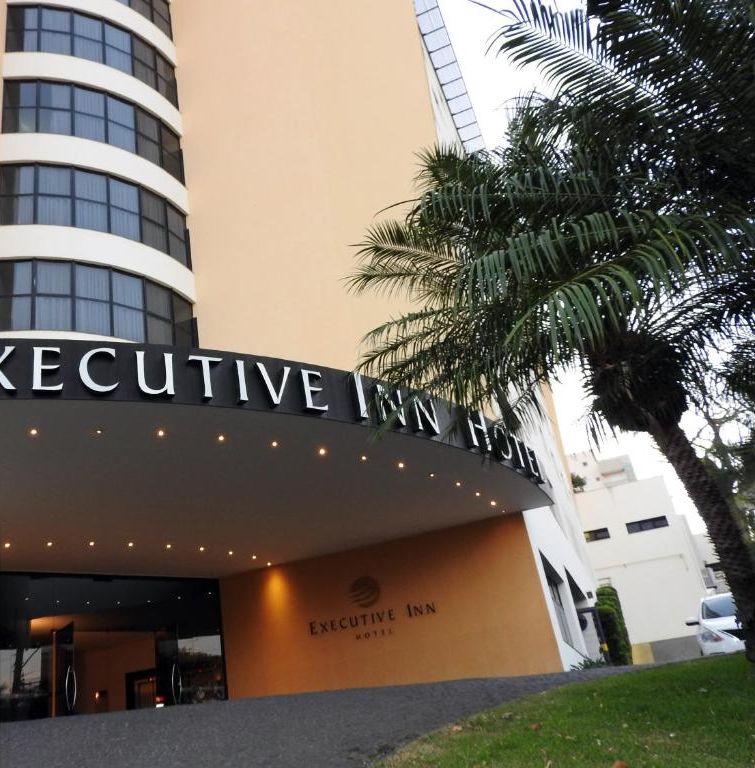 Exterior do Executive Inn Hotel, com uma placa visível e o logotipo do TripAdvisor indicando um prêmio Travelers' Choice de 2020.