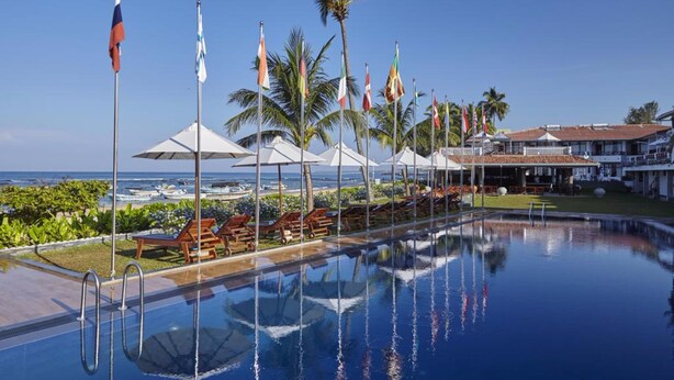 A pool area with lounge chairs and umbrellas, flags, and a view of the ocean. - Coral Sands Hotel (Hikkaduwa)