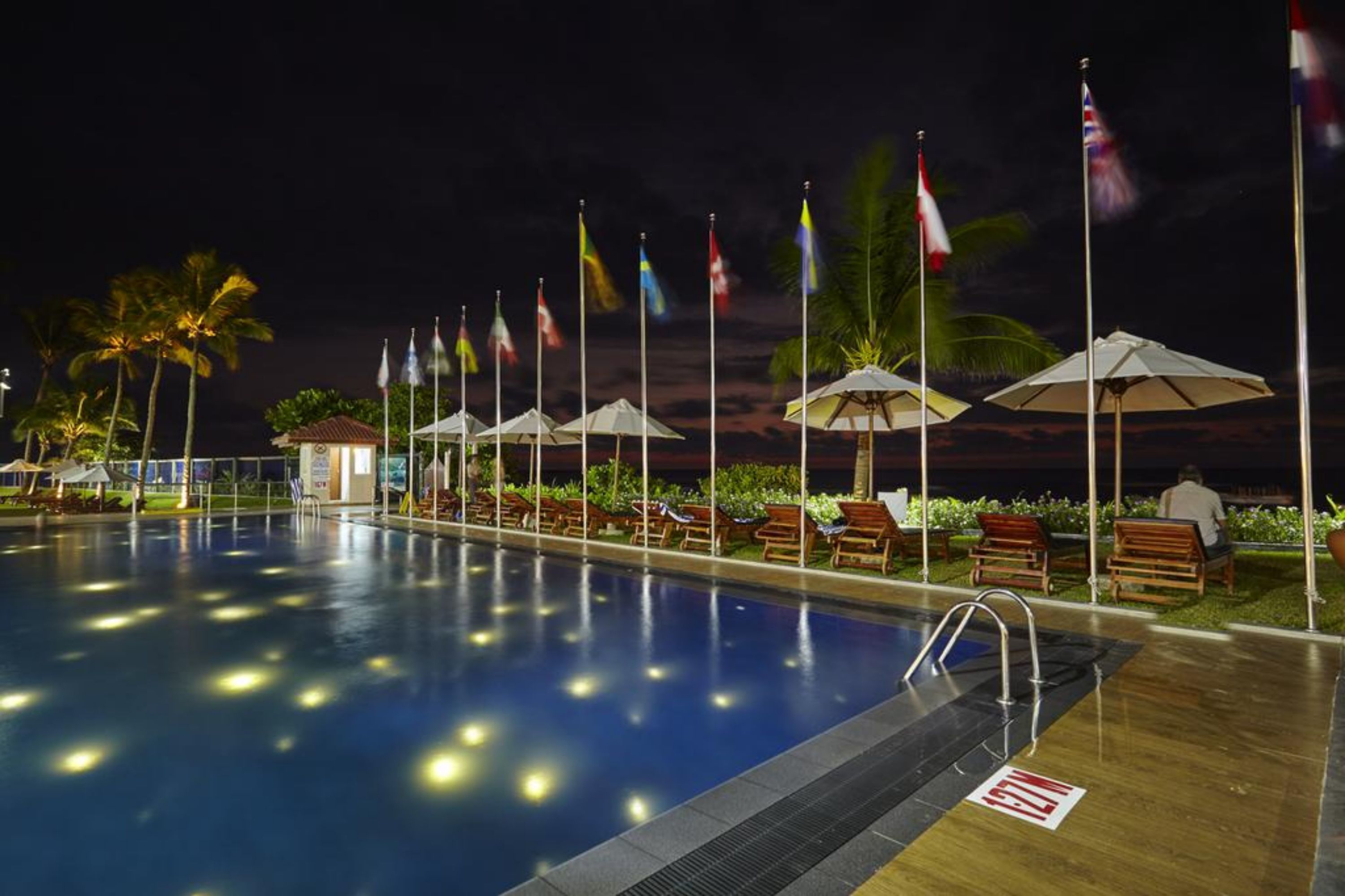 A swimming pool with lounge chairs and umbrellas, surrounded by flags and palm trees.
