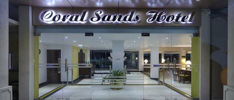 De ingang van Hotel Coral Sands met een glazen gevel, een receptie en een zichtbare kamerplant.
