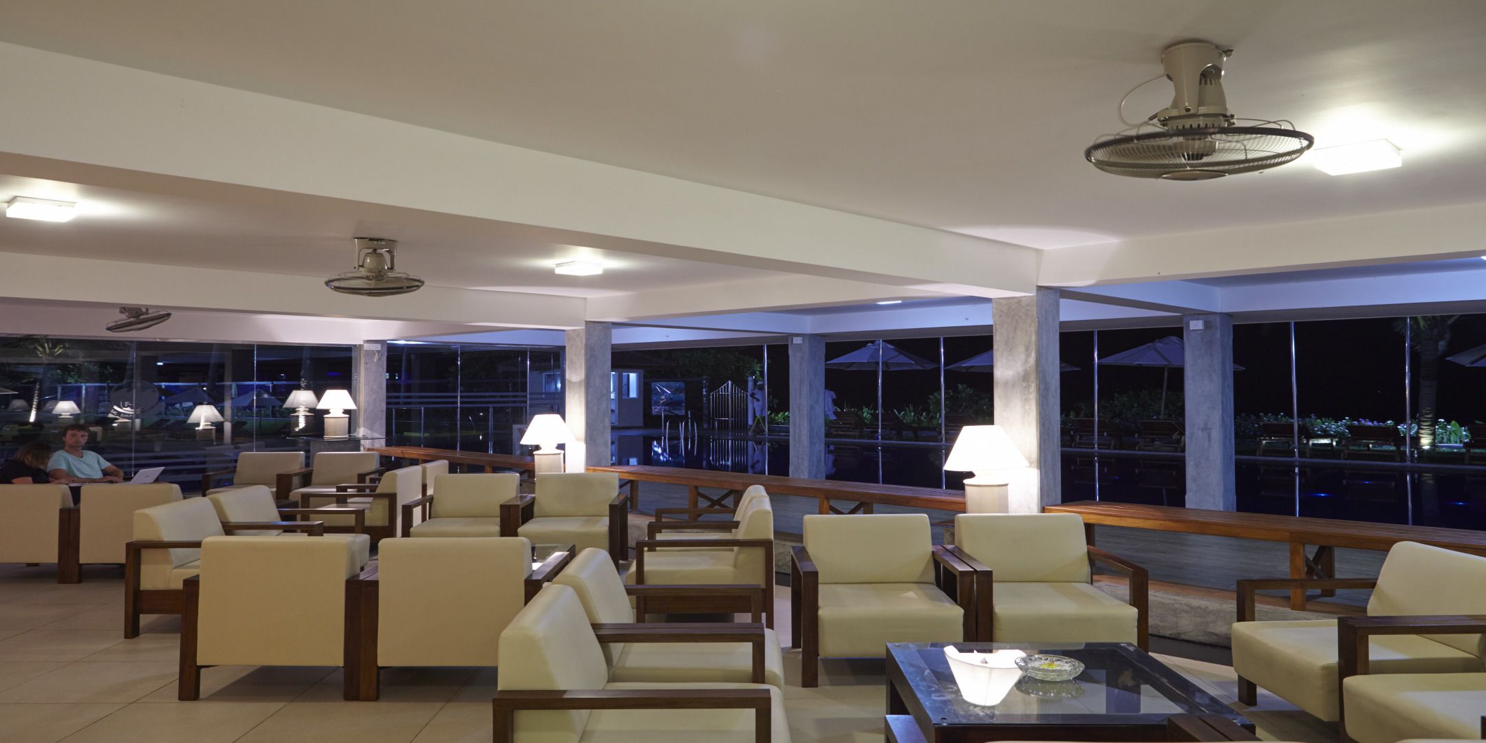 Amplia sala de estar con diversas opciones de asientos, ventanas grandes que ofrecen vistas del cielo nocturno y una piscina, además de ventiladores de techo para ventilación.