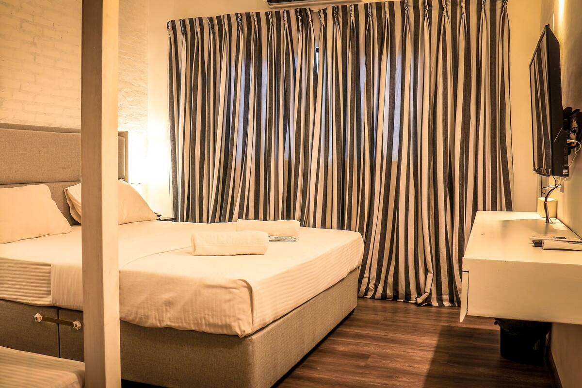 A hotel room with a bed, a bedside table, a mirror, and a window with striped curtains.