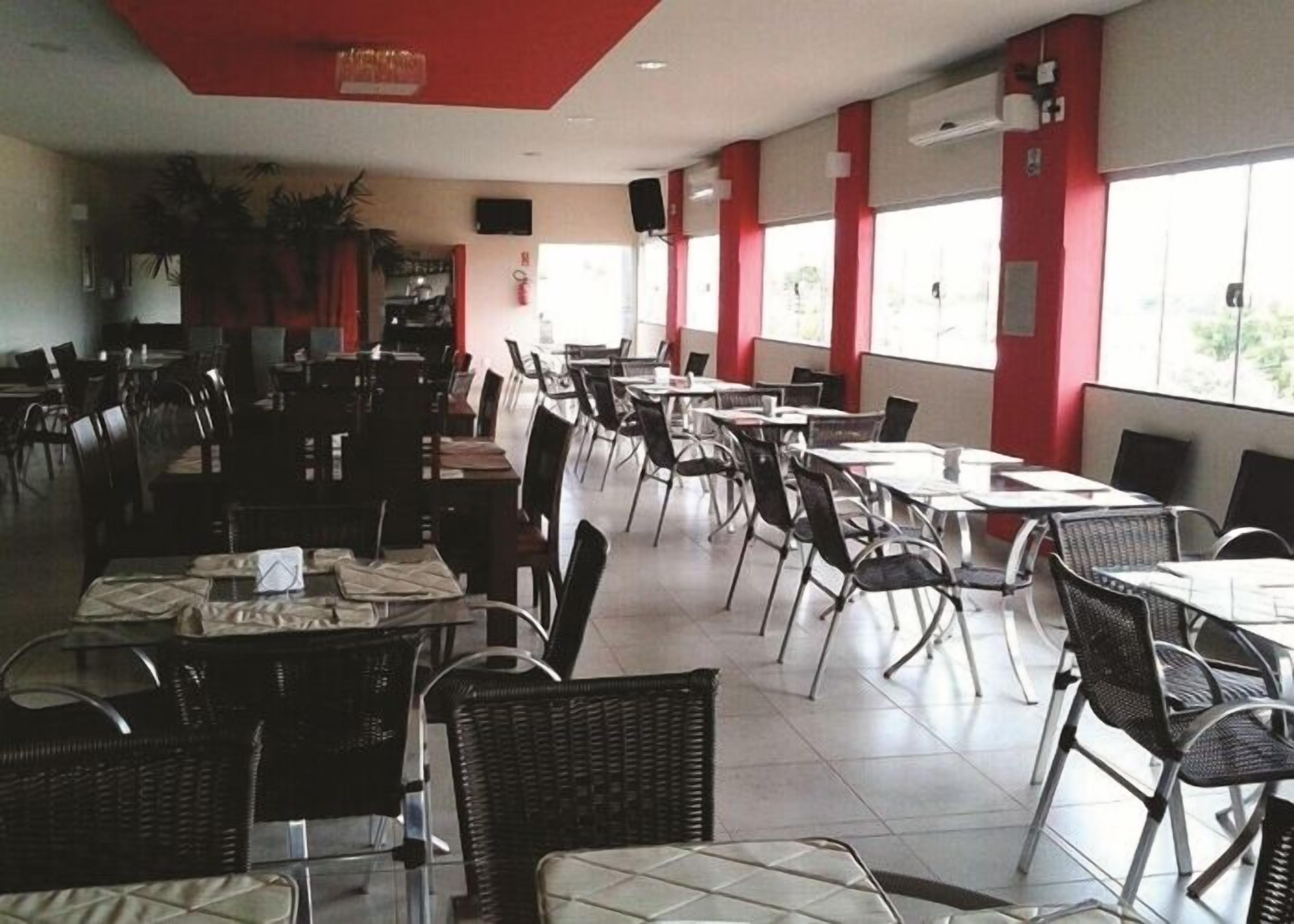 A spacious restaurant with high ceilings, red accents, and large windows allowing natural light. Tables and chairs are neatly arranged, and the floor is tiled.